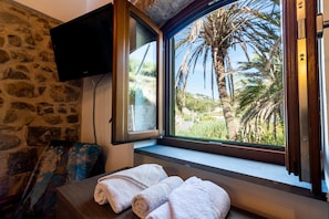A room with a stone wall, a window with a view of palm trees, a flat-screen TV, and two towels on a wooden bench. - Monet House surrounded by nature and the sea (Bordighera)