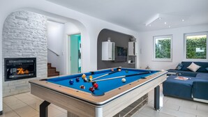 Una mesa de billar en un salón moderno con chimenea de piedra, un sofá y ventanas amplias.