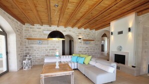 Ein geräumiges Wohnzimmer mit Steinwand, Holzbalkendecke, Kamin und einer großen Couch mit bunten Kissen.