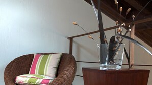 Une chaise en osier avec un coussin à rayures, une table sur pied en bois surmontée d’un vase en verre contenant des brindilles et des fleurs séchées, et un plancher en bois.