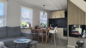 Ein modernes Wohnzimmer mit einem grauen Sofa, einem Esstisch mit Stühlen, einem kleinen runden Tisch und einer Küchenzeile mit Holzausbau, einem Herd und einem Grill.