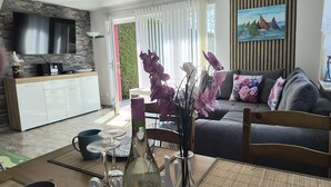 Ein modernes Wohnzimmer mit einer Couch, einem Esstisch mit Blumenvase, einer Küchenzeile mit Fernseher und einem Bild an der Wand.