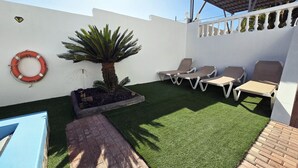 Un patio con césped artificial, una palmera, sillas de descanso y un flotador.