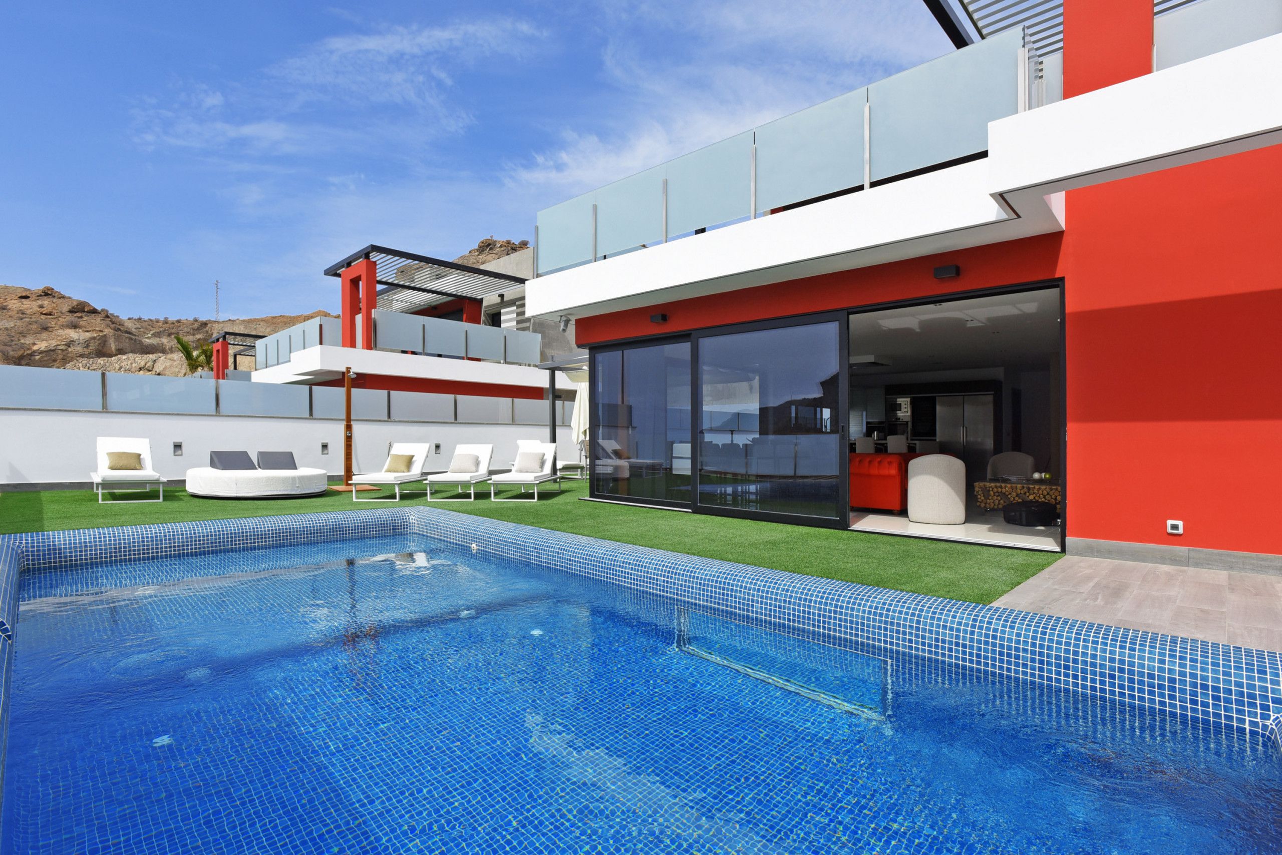 A modern pool area with a clear blue pool, white lounge chairs, and a red and white building with large glass windows.
