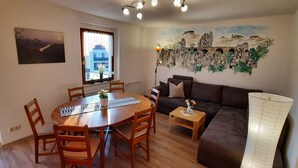 A cozy living room with a wooden dining table, a sofa, and a large window with a view of a building. - Apartment with garden and pool use (Rathmannsdorf)
