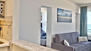 Une chambre d’hôtel moderne avec un canapé gris, un tableau représentant un paysage et une vue sur la mer par la fenêtre.