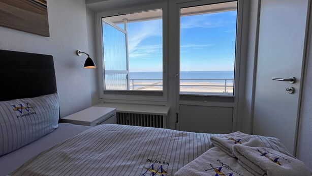 Une chambre à coucher comprenant un lit, une table de chevet, une lampe, une fenêtre donnant sur la plage et une porte.