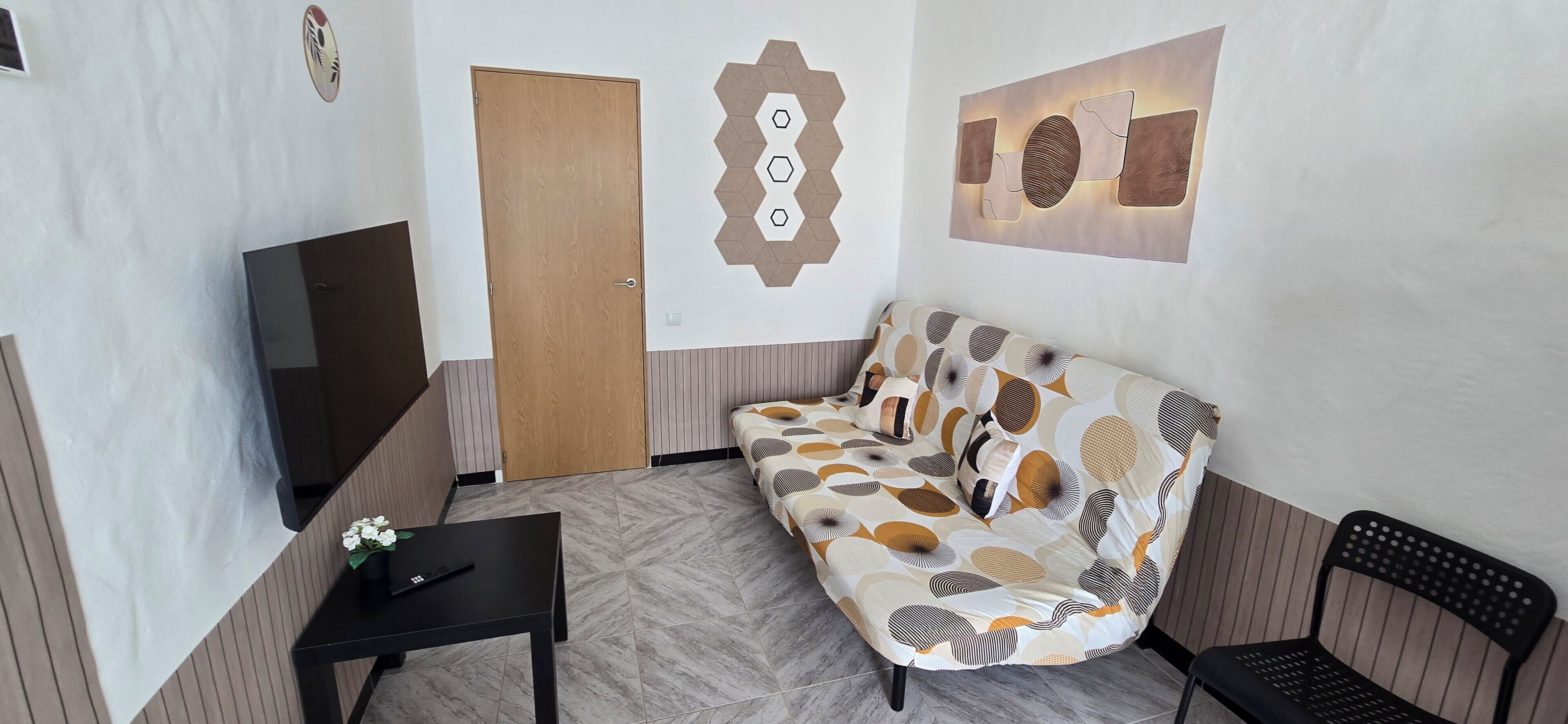 A room with a sofa, a small table, a chair, a wooden door, and a wall with geometric patterns.