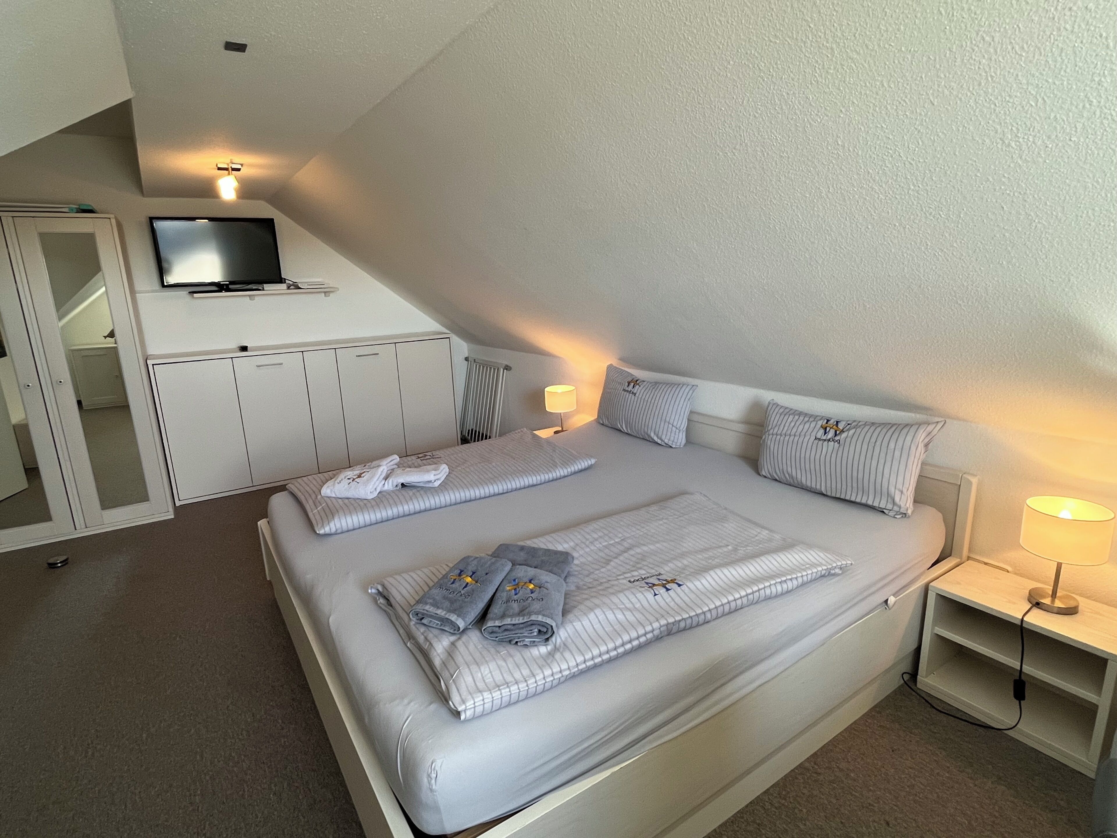 A bedroom with a bed, a TV mounted on the wall, a bedside table with a lamp, and a wardrobe.