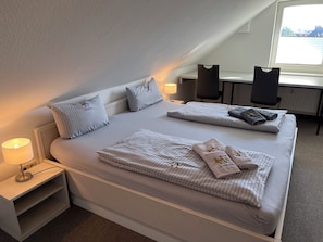 A hotel room with a bed, two pillows, a bedside lamp, a desk, and a chair.