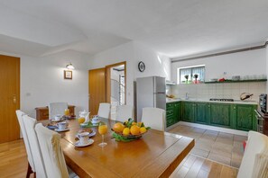A dining area with a wooden table set for breakfast, a kitchen with green cabinets, and a window with plants.