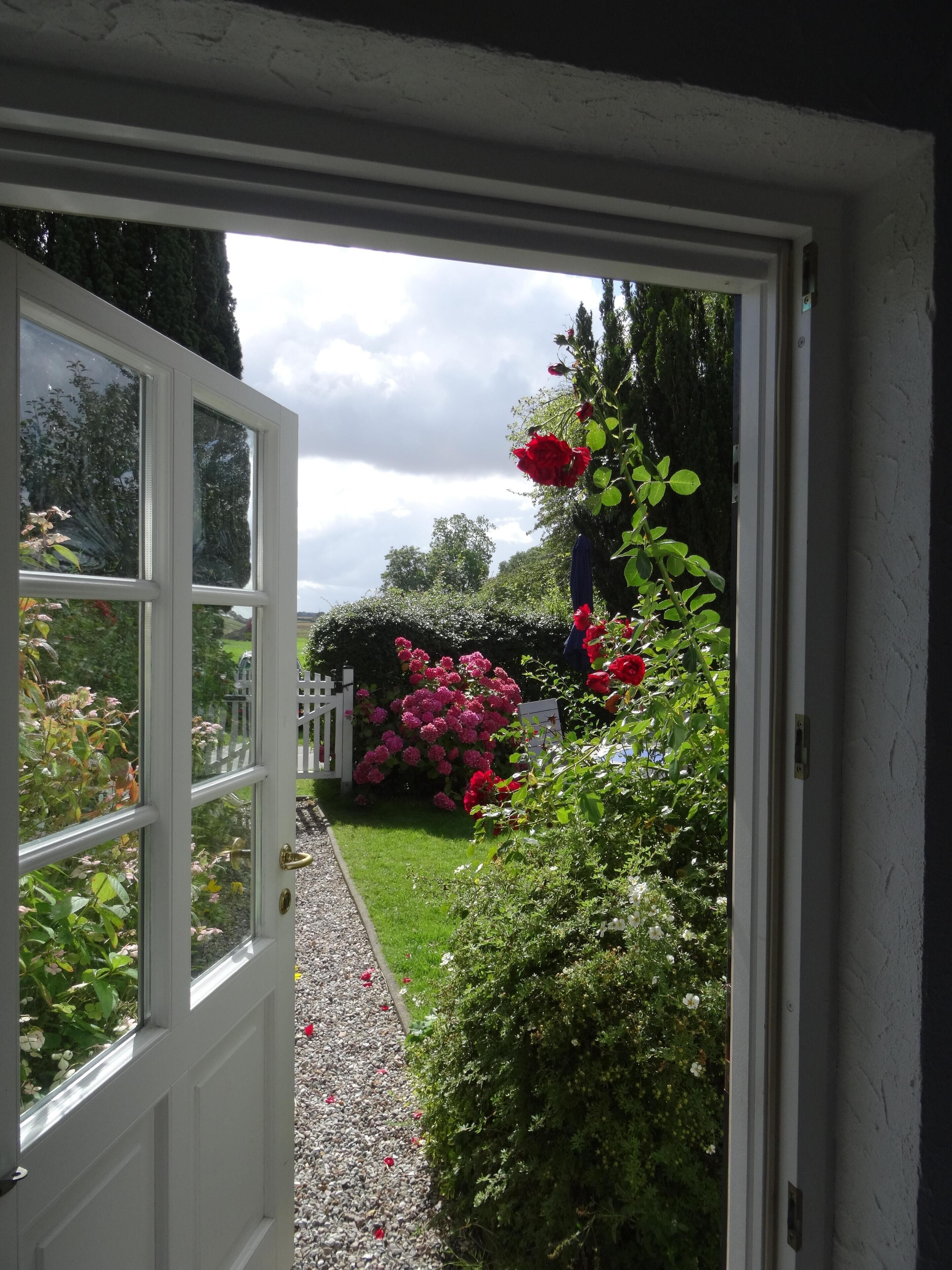 Une porte blanche donnant sur un jardin avec un chemin de gravier, des fleurs roses et rouges, et une clôture blanche en bois.