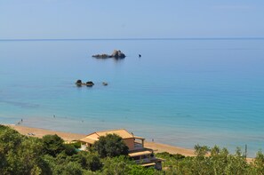 Vista costera con playa, aguas azules cristalinas y un edificio con techo de tejas.