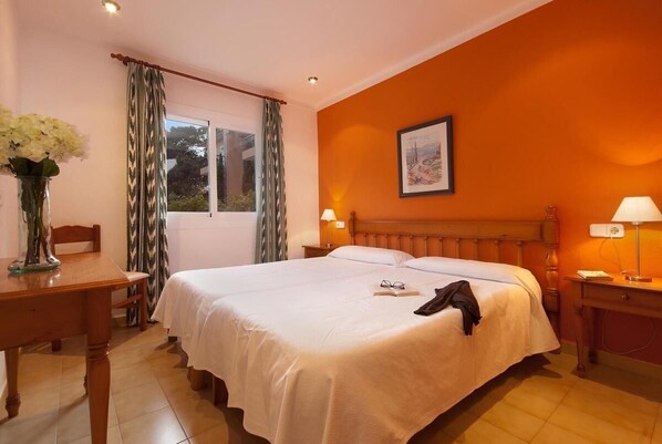 A hotel room with a large bed, a wooden headboard, a bedside table with a lamp, a window with curtains, and a framed picture on the wall.