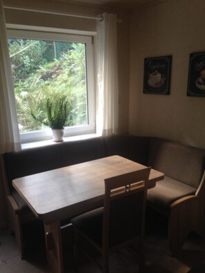 Uma sala de jantar com mesa e cadeiras de madeira, um banco acolchoado, uma janela com uma planta em vaso e duas imagens emolduradas na parede.