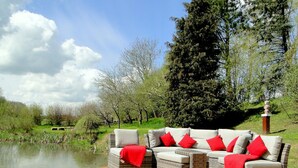 A wicker sofa set with red cushions on a wooden deck by a lake, surrounded by trees and greenery. - "NATÜRlich Wassermühle" Vacation Home in Kappeln (Kappeln)