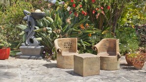 Een tuin met een stenen beeld, twee houten stoelen en een bijzettafeltje.