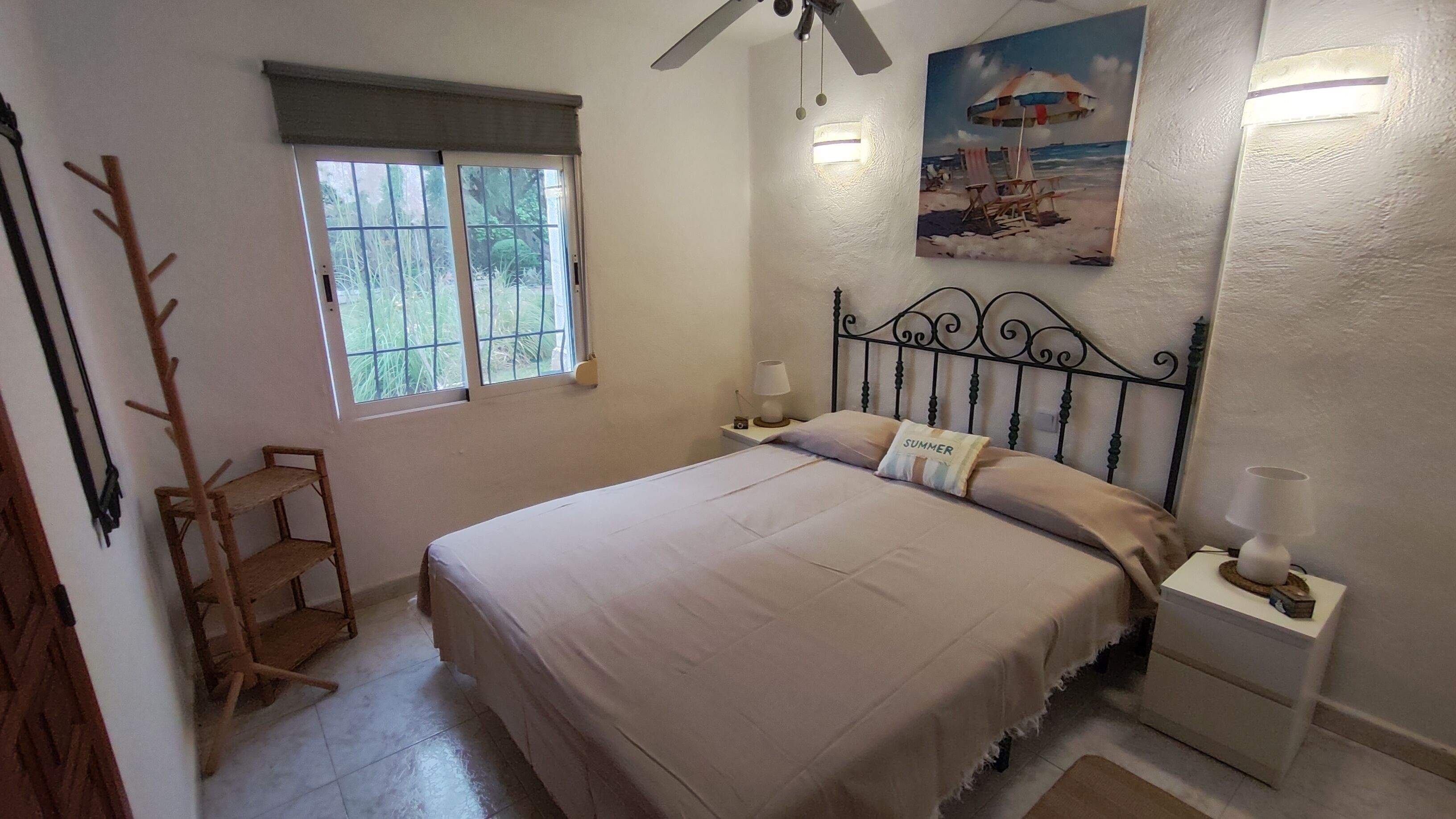 A bedroom with a bed, a nightstand, a lamp, a window with a view of greenery, and a ceiling fan.