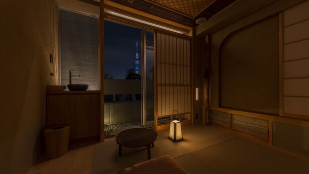 Una habitación japonesa tradicional con suelo de tatami, una puerta corredera, una pequeña mesa, un farol y vistas de un paisaje urbano con una torre.