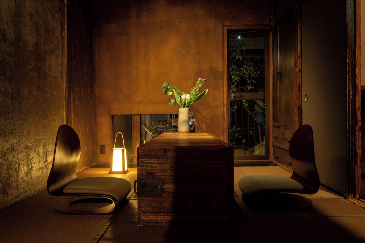 A modern room with a wooden table, two chairs, a vase with flowers, and a lit lamp.