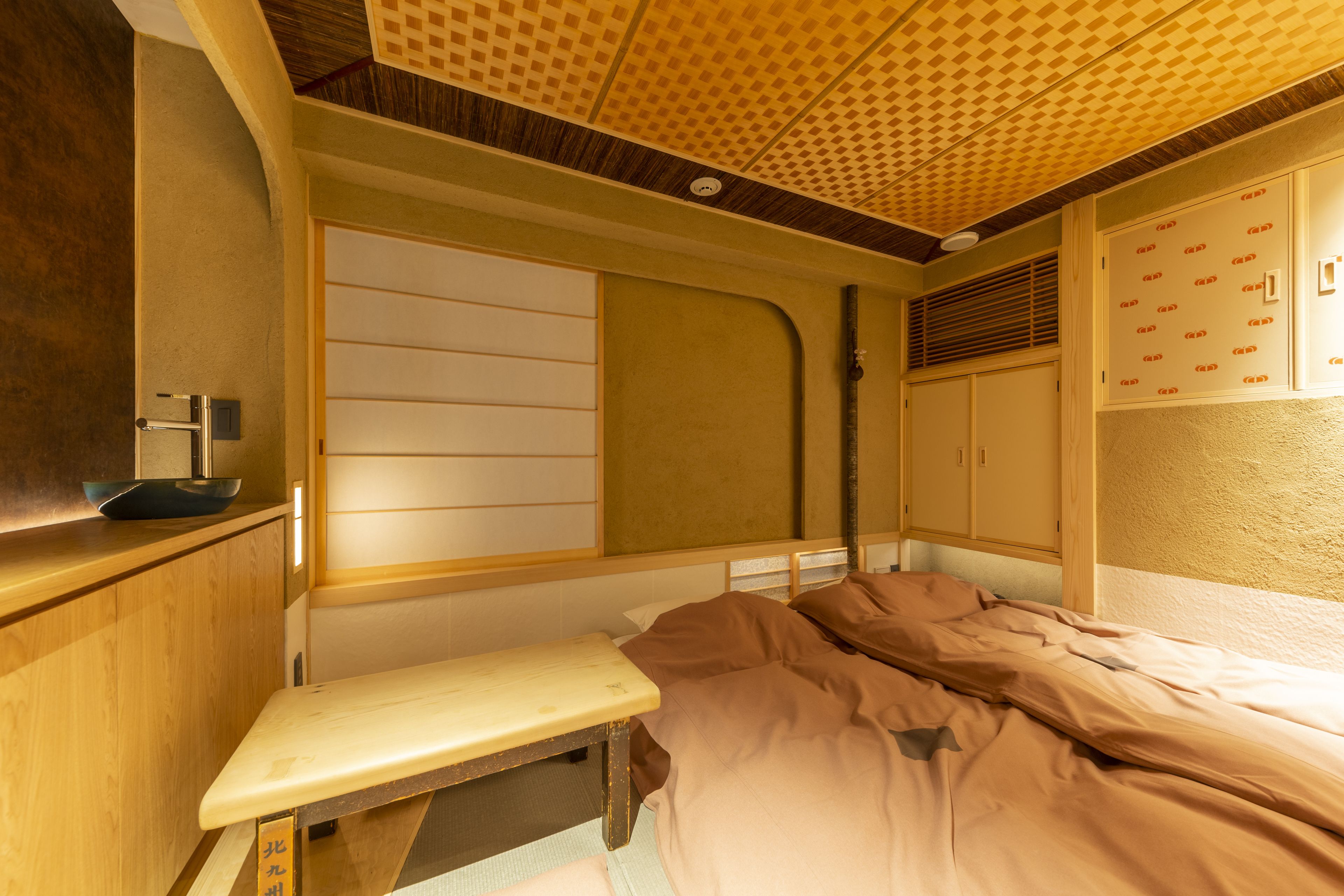 A room with a bed, a bench, a wooden cabinet, and a wall with a patterned design.