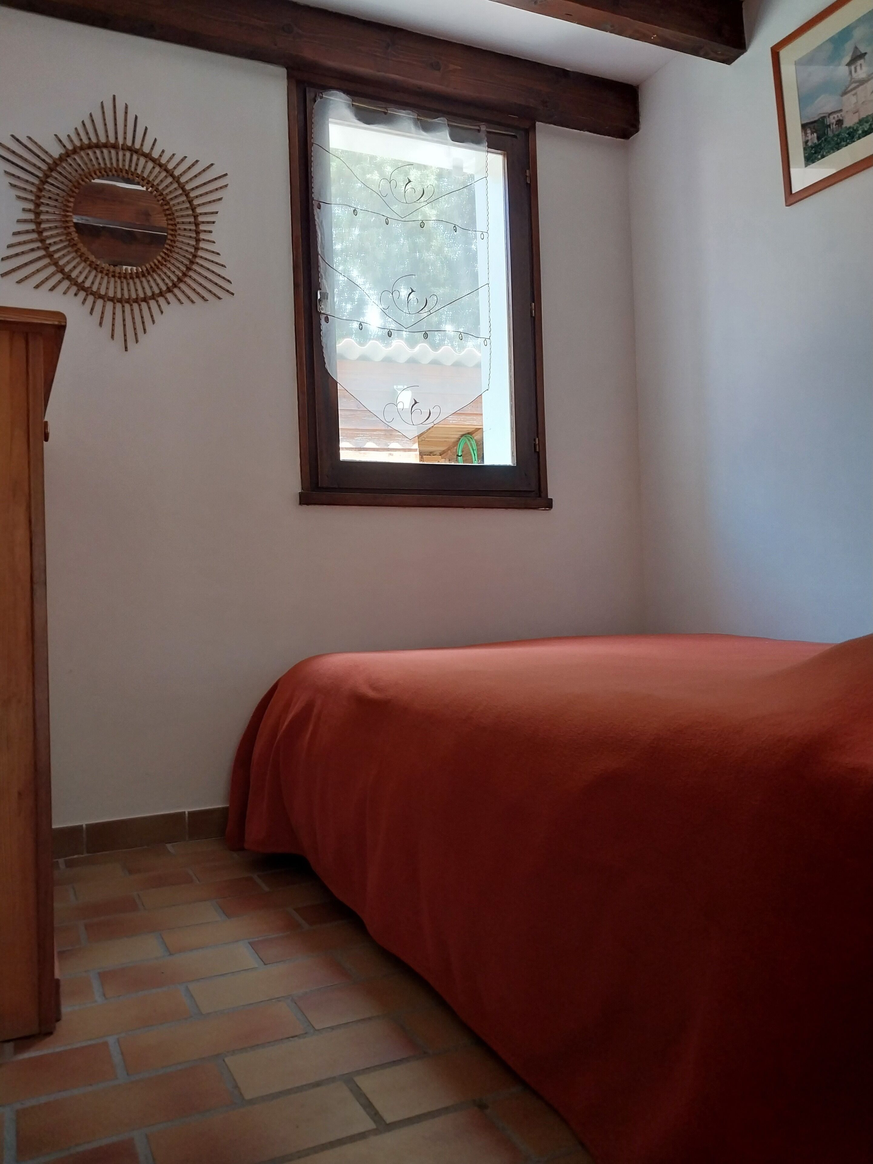 A bedroom with a bed, a window with decorative curtains, a wooden headboard, and a wall-mounted mirror.