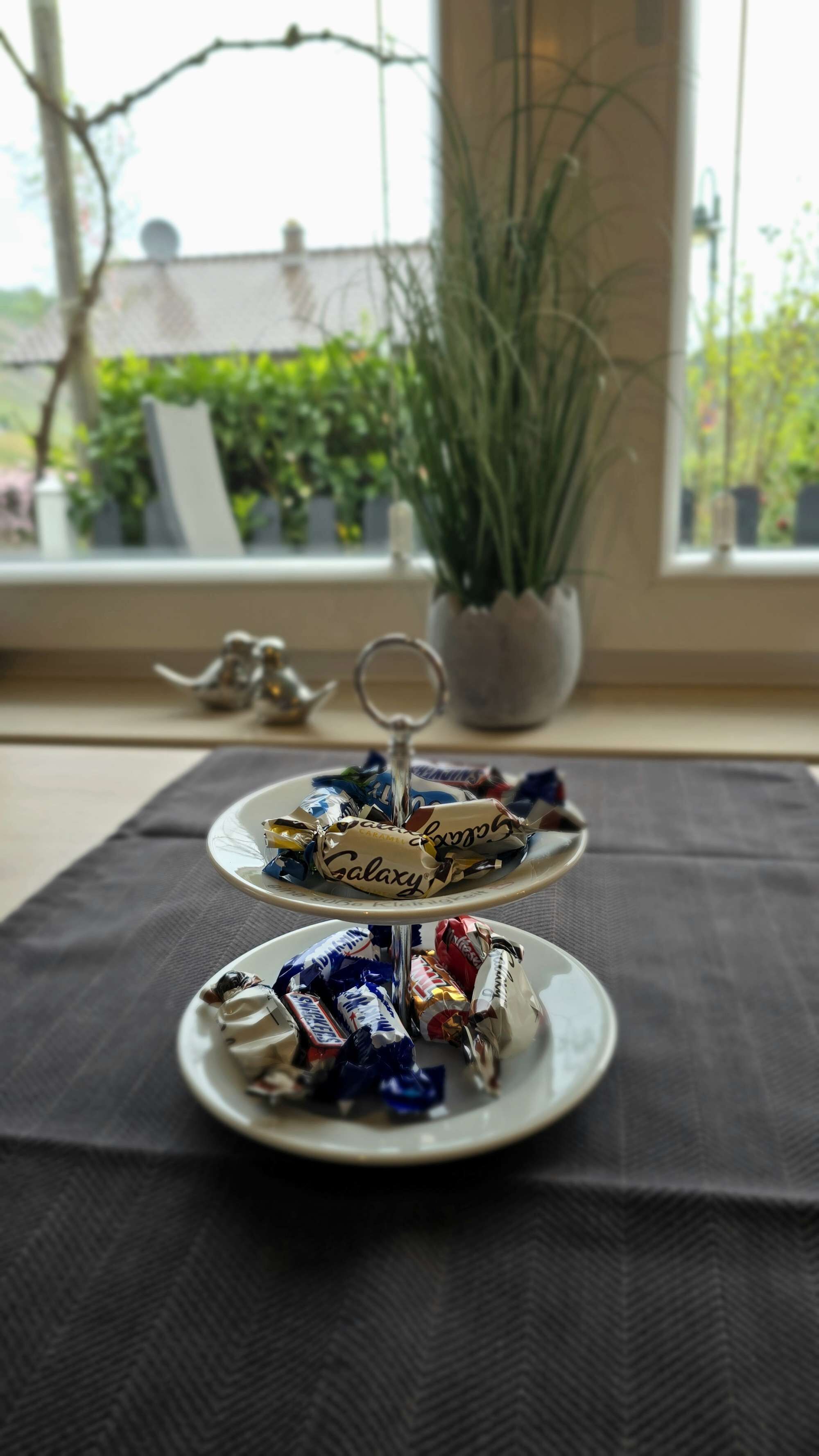 Ett våningsfat med olika sorters godis står på ett bord nära ett fönster med utsikt över grönskan utanför.