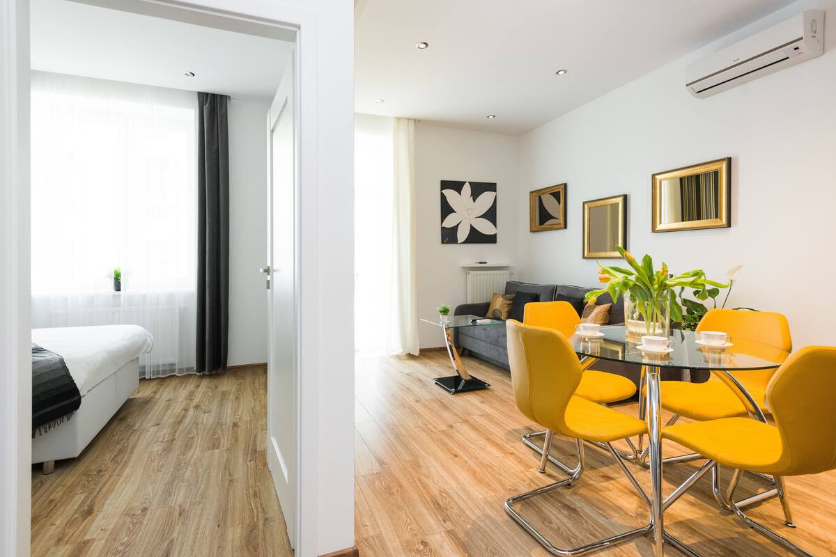 Ein modernes Hotelzimmer mit einem Bett, einem Glastisch mit gelben Stühlen und einer Essecke mit einer Couch und einer Pflanze.