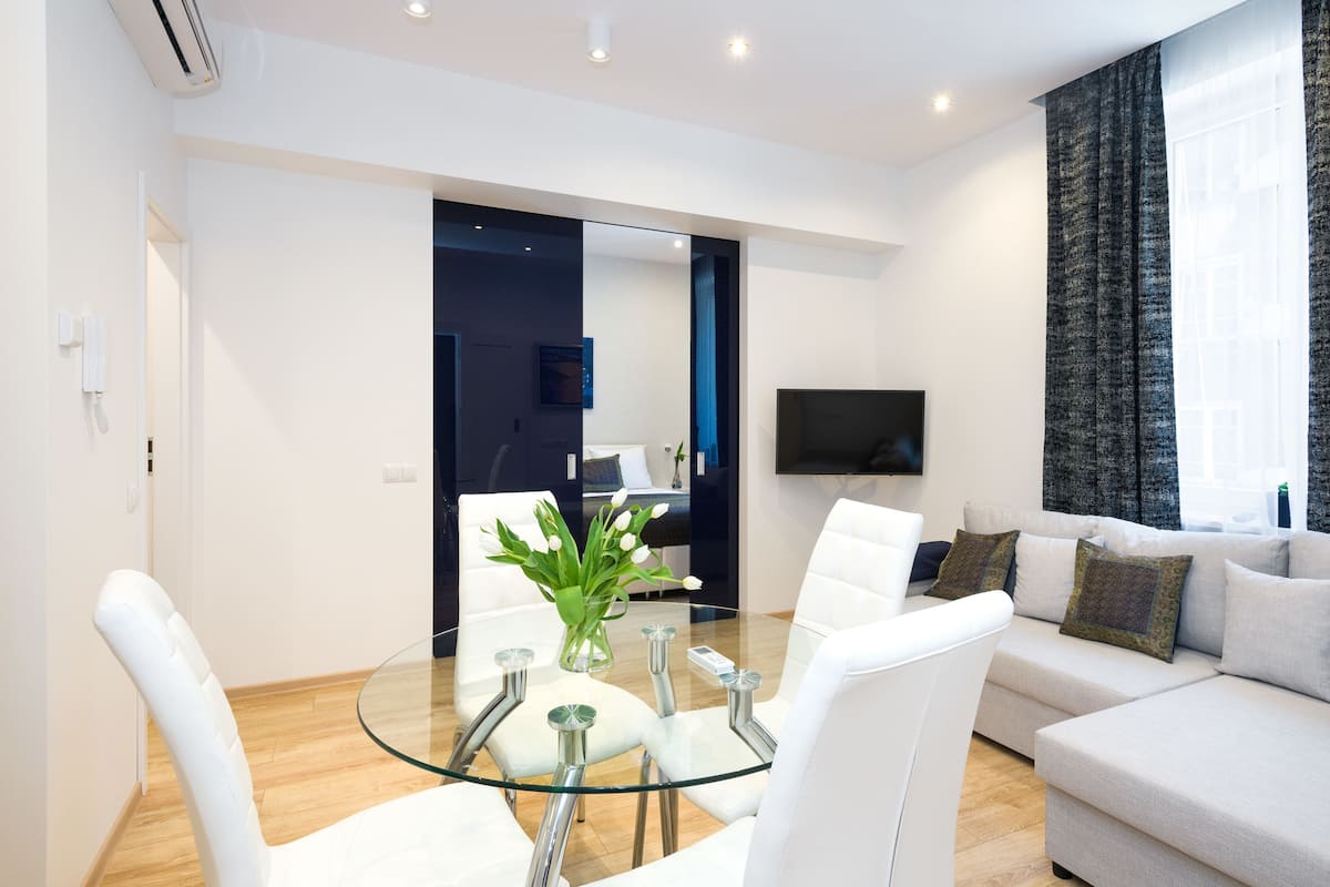 Ein modernes Wohnzimmer mit einem Glastisch, weißen Stühlen, einem grauen Sofa und einem an der Wand montierten Flachbildfernseher.