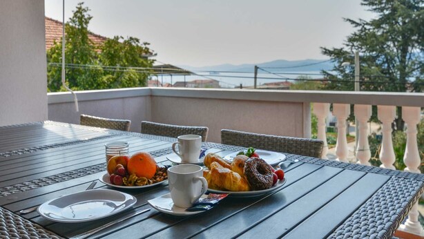 A balcony table with breakfast items, including fruits, pastries, and a cup, overlooking a scenic view with trees and mountains.