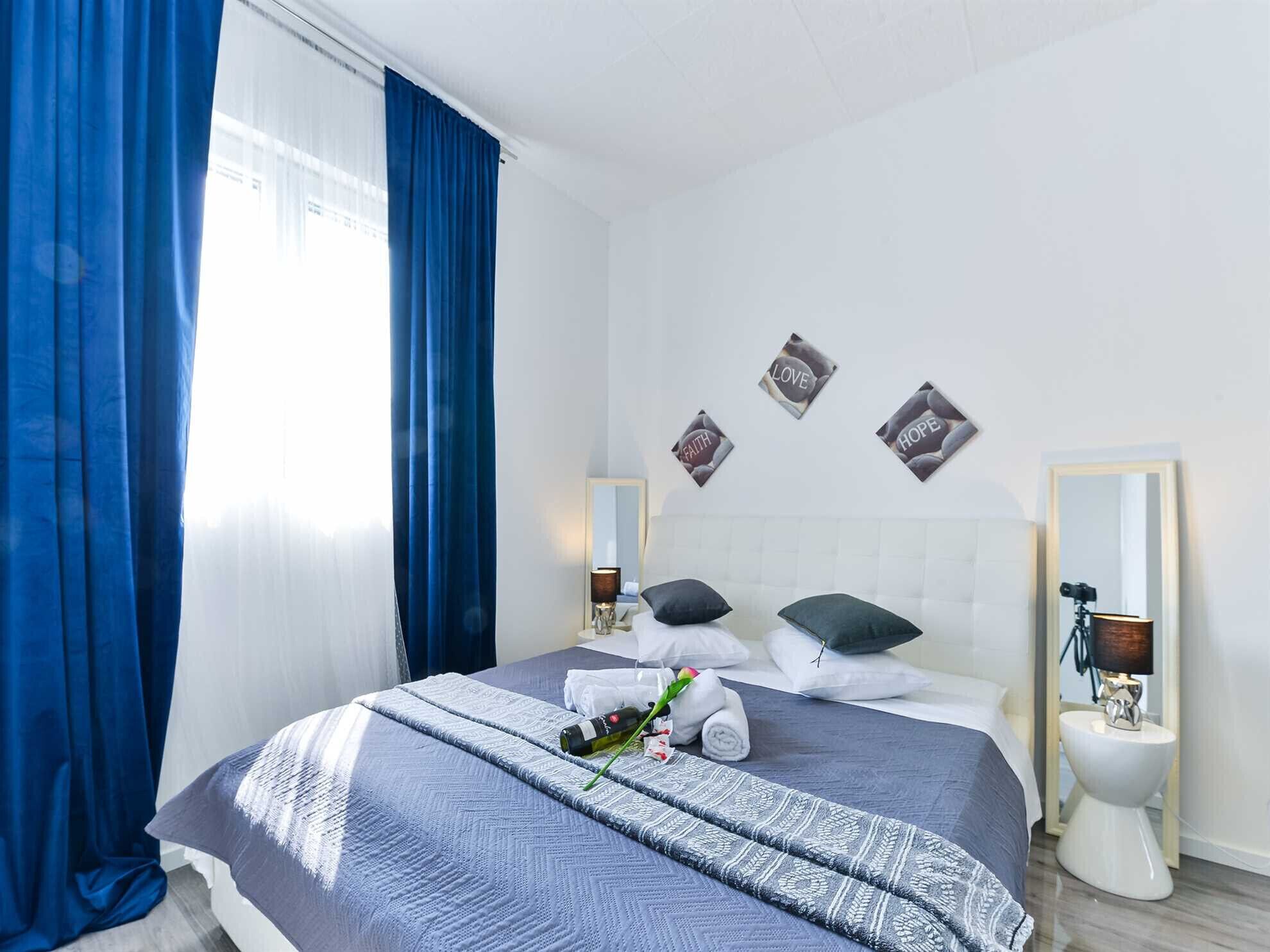 Ett hotellrum med en säng, ett fönster med blå gardiner, ett sängbord med en lampa och en spegel.