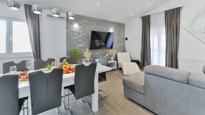 A modern living room with a grey sofa, a dining area with a table set for four, and a mounted flat-screen TV on a stone wall.