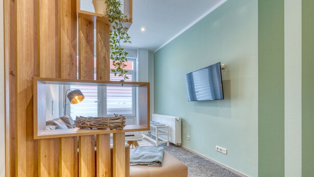 Una habitación con un separador de madera, una cama, un televisor montado en la pared y una ventana con persianas.