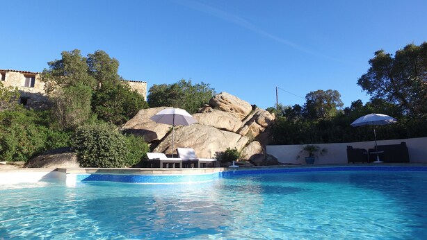 Piscina con sillas de descanso y sombrillas, rodeada de rocas grandes y follaje.