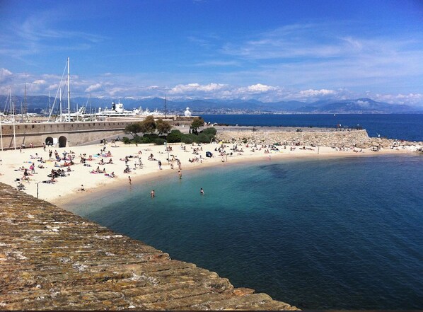 A coastal beach with a stone wall, clear blue water, and boats in the background.