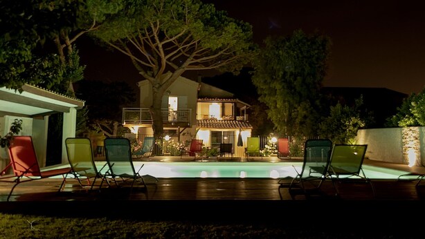 Un espace aménagé au bord de la piscine, avec des chaises longues, une terrasse en bois et une maison dotée de plusieurs balcons et d’un éclairage extérieur.