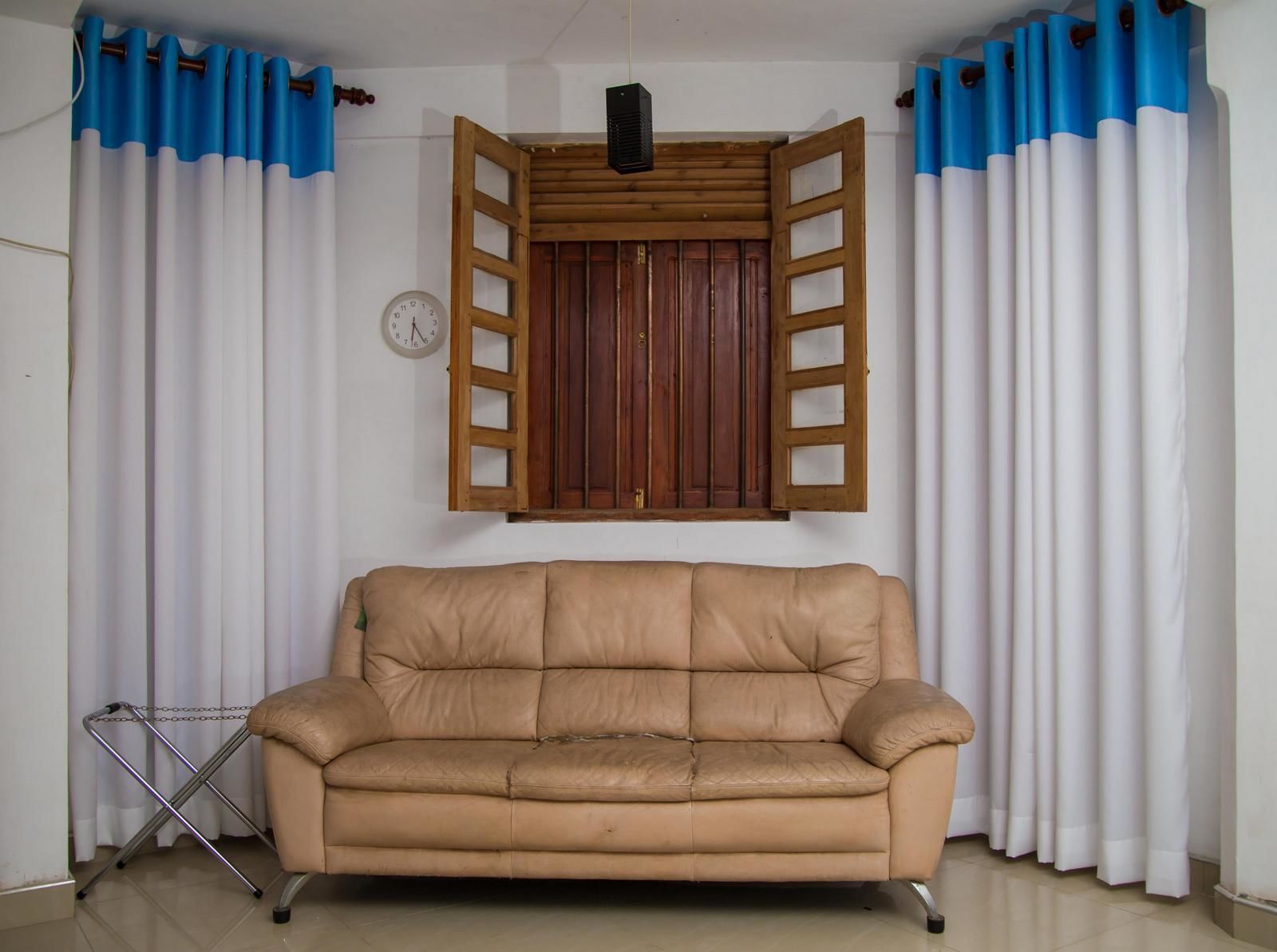 A tan leather sofa in a room with white and blue curtains, a wooden window shutter, and a clock on the wall.