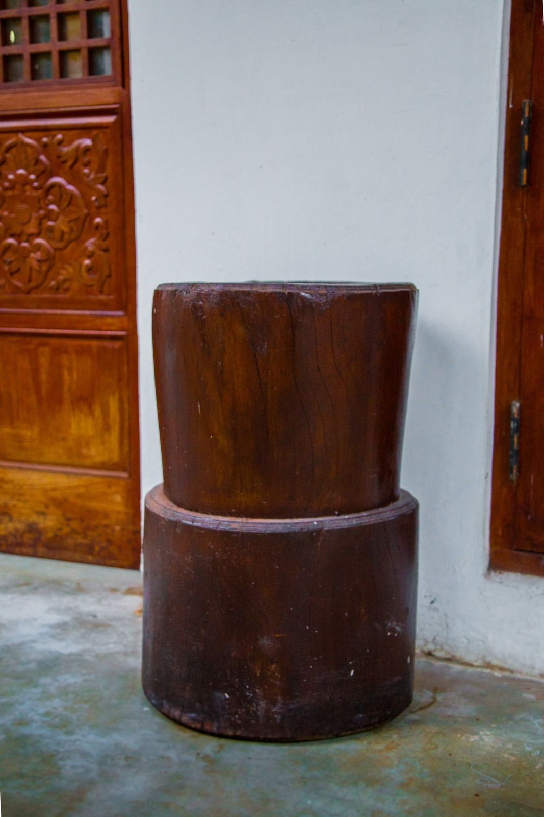 A wooden stool with a carved design, placed on a concrete floor against a white wall with a wooden door in the background.