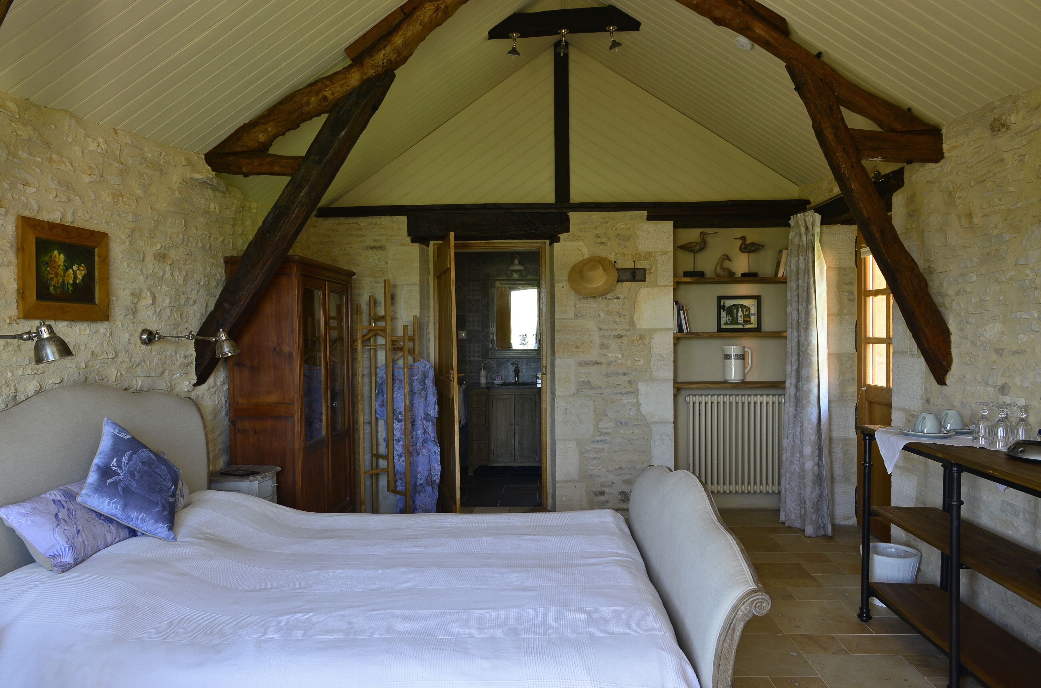 Une chambre à coucher avec un grand lit, des poutres en bois, des murs en pierre, une porte en bois et une petite table avec des verres.