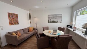 A modern living room with a sofa, chairs, a round table, artwork on the walls, and a window with blinds. - Maike - Apartment with terrace and small garden (Wangerooge)