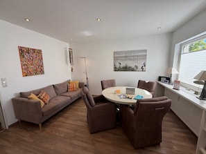 A modern living room with a sofa, chairs, a round table, artwork on the walls, and a window with blinds.