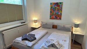 A neatly made bed with striped bedding and towels, two bedside tables with lamps, a radiator, and a colorful abstract painting on the wall. - Maike - Apartment with terrace and small garden (Wangerooge)