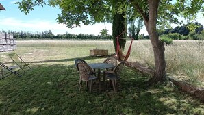 An outdoor seating area with a table and chairs under a tree, surrounded by a grassy field and a wooden structure in the background.