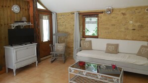 Ein gemütliches Zimmer mit Holzbalkendecke, einem weißen Sofa mit Kissen in Herzmuster, einem Glastisch, einer weißen TV-Konsole und einem Holzschaukelstuhl.