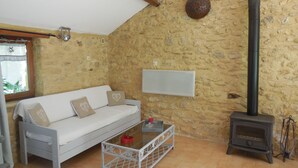 Ein gemütlicher Raum mit Steinwand, einem weißen Sofa mit gemusterten Kissen, einem Glastisch, einer Whiteboard und einem Kaminofen.