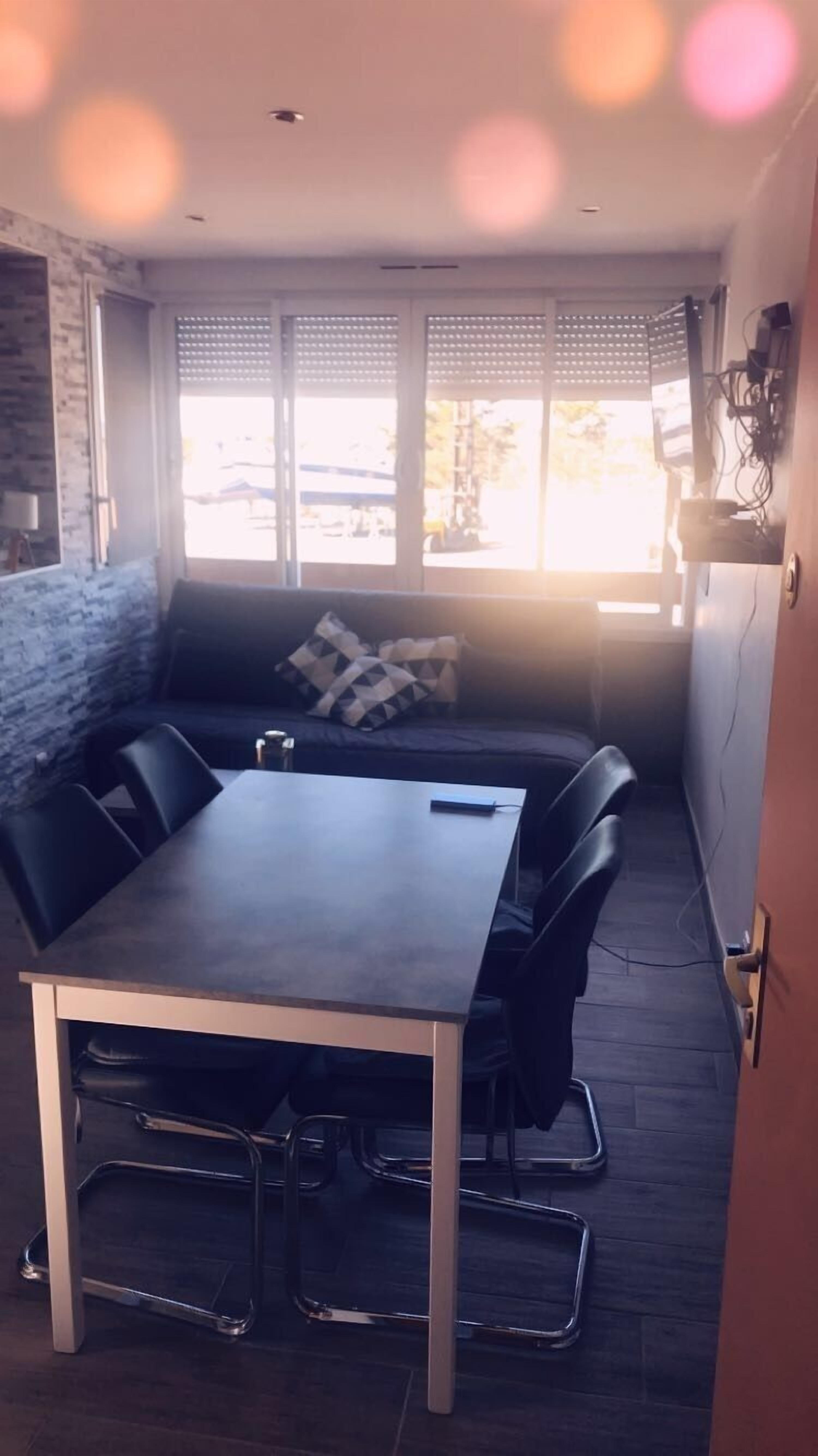 A dining area with a table and chairs, a sofa with cushions, and a view through large windows.