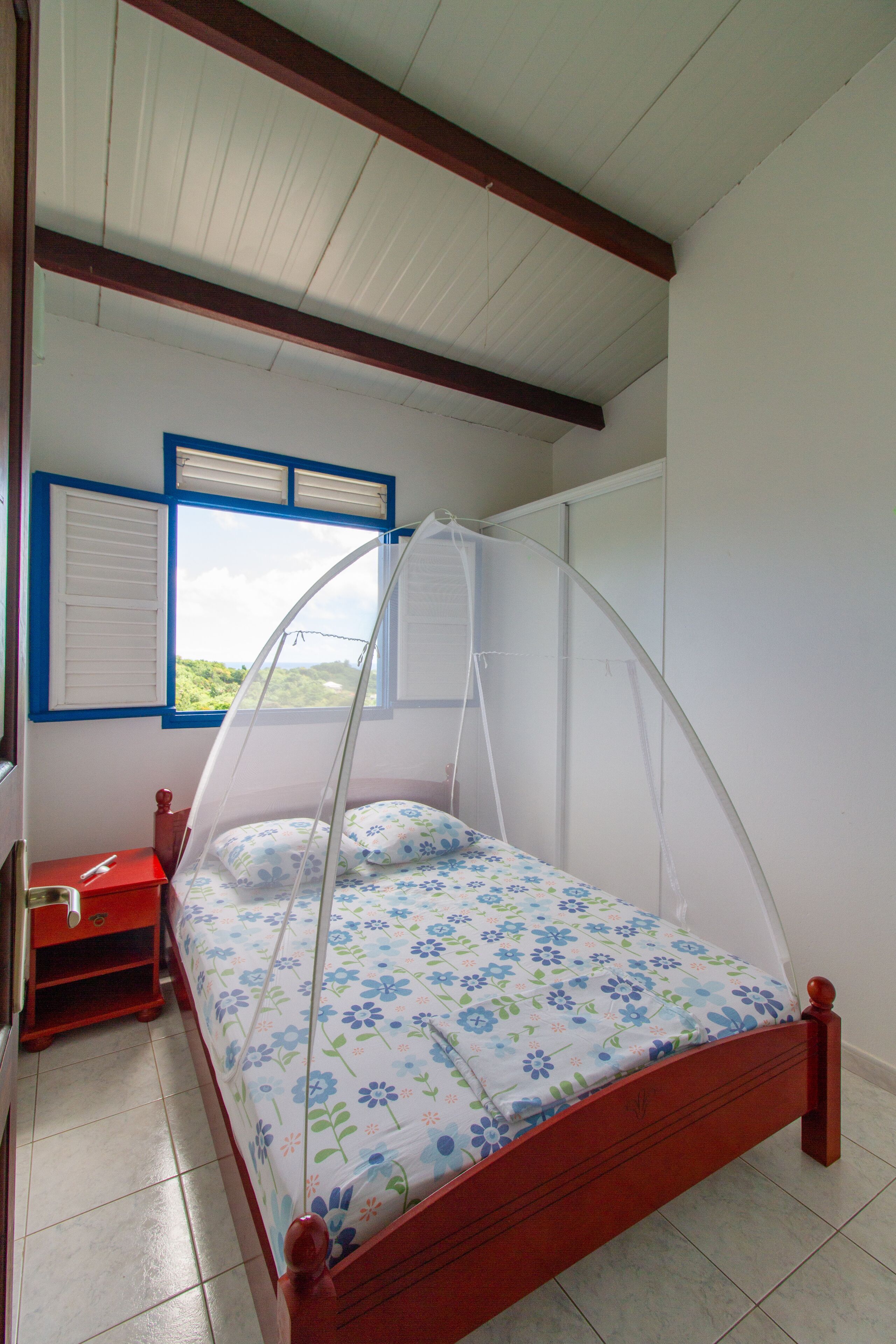 Une chambre à coucher avec un lit, une fenêtre avec des volets, une table de chevet rouge et un plafond en bois.