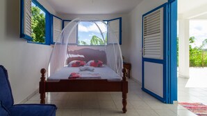 A bedroom with a bed under a mosquito net, a chair, and a view of the outdoors through open shutters. - Villa between sea and countryside (Le Marin)