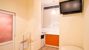 Una habitación compacta con suelo de baldosas, una mesa blanca con dos taburetes naranjas, un televisor montado en la pared y una ventana con persianas.