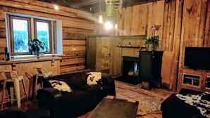 Interior rústico de una cabaña con paredes de madera, una chimenea, un televisor y una ventana con vistas a árboles.